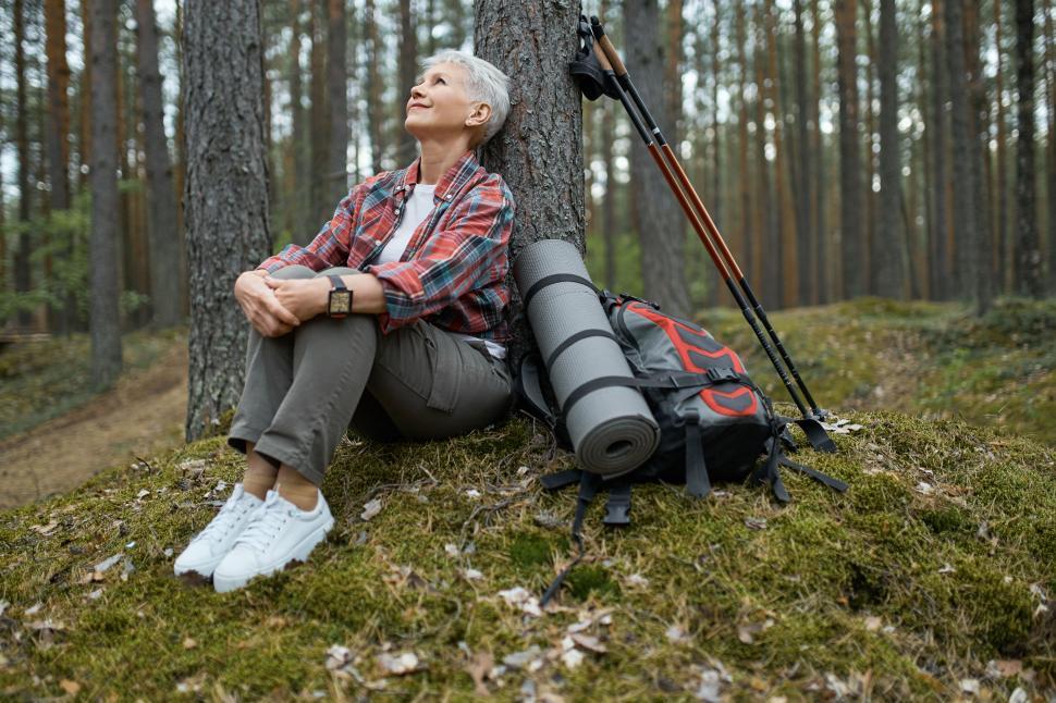 Free Image of Beautiful mature woman in sneakers and activewear sitting on grass under pine having rest during nordic walking with sticks and backpack, looking up with relaxed carefree smile, breathing fresh air 