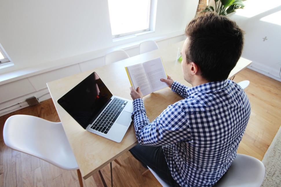 Free Image of Man reading book at wooden table with laptop beside. 