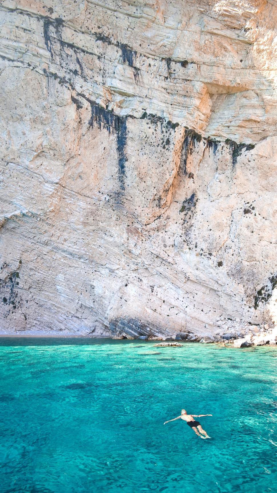 Free Image of Person floating in clear turquoise water by a cliff face. 
