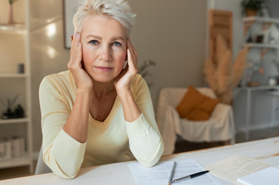 Free Image of Portrait of sad middle aged female journalist with short white hair sitting at desk with papers, holding hands on her head, feeling tired, trying to focus on work. People, age and occupation 