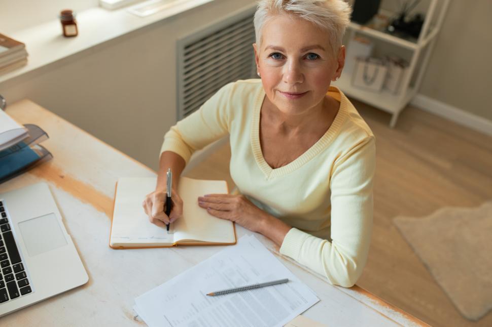 Free Image of Top view of successful 50 year old female entrepreneur sitting at desk with laptop and papers, writing down ideas in copybook, looking at camera with confident smile. People, occupation and technology 