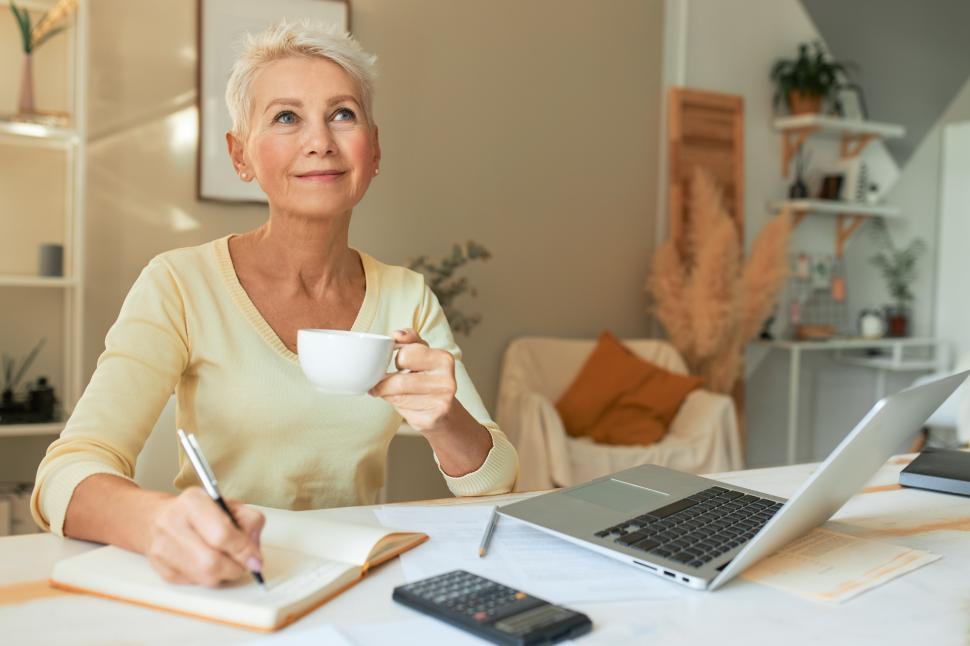 Free Image of Job, occupation and technology concept. Portrait of self employed mature female sitting at desk at home with laptop, calculator, holding cup and pen, writing ideas in diary, having pensive look 