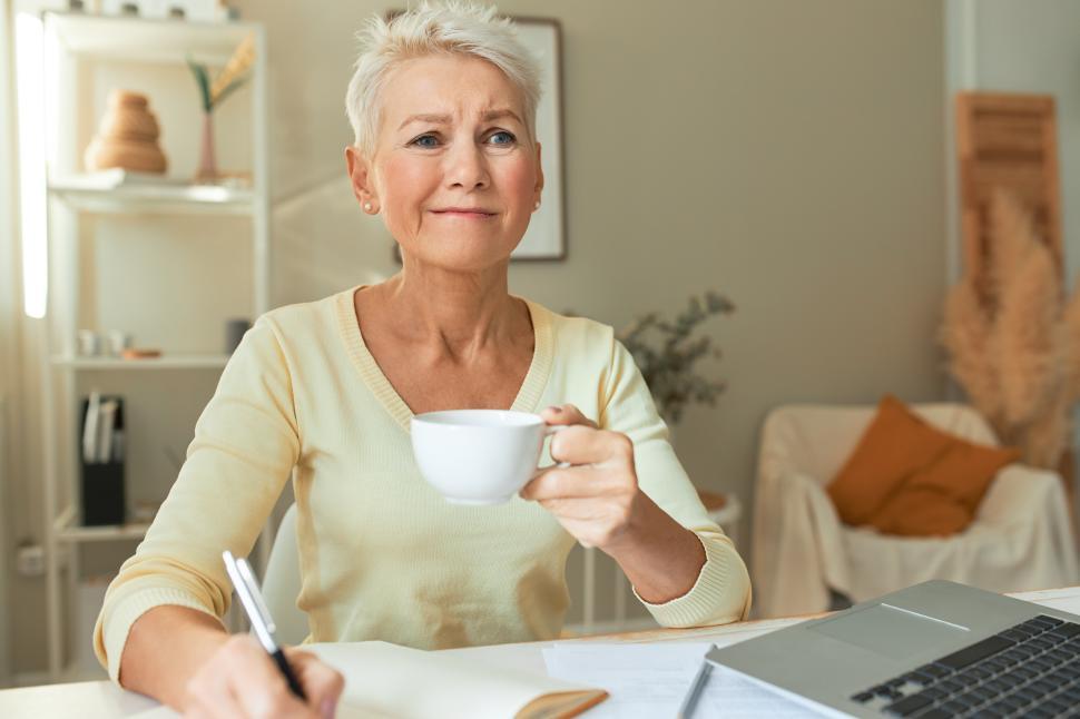 Free Image of Horizontal image of sad nervous mature female entrepreneur using portable computer, holding mug, handwriting in daily planner, having frustrated facial expression because of too much work to do 