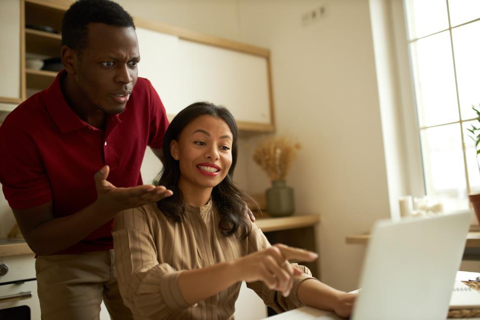 Free Image of Dark-skinned man standing next to his beautiful young female business partner sitting at kitchen table, in front of opened laptop, pointing at screen, solving problem with wireless connection 