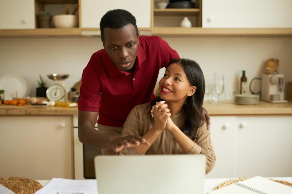 Free Image of Pretty happy mixed-race woman trade investor sitting in front of laptop against kitchen background with her husband standing behind her looking at screen with shocked face expression 