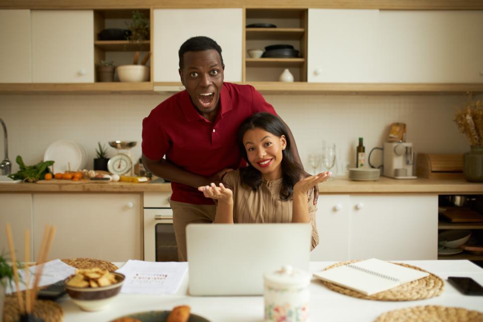 Free Image of Happy ethnicity couple. Cheerful super excited African guy in red t-shirt standing behind his successful shrugging smiling wife sitting at table in front of laptop, celebrating business start up 