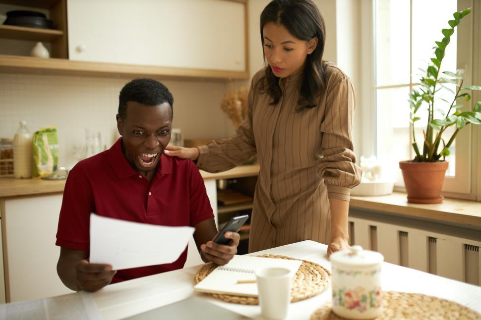 Free Image of Pretty mixed-race female in brown casual dress calming down her frustrated husband screaming with anger and despair holding tax bill and mobile phone, touching his shoulder, sitting at kitchen 