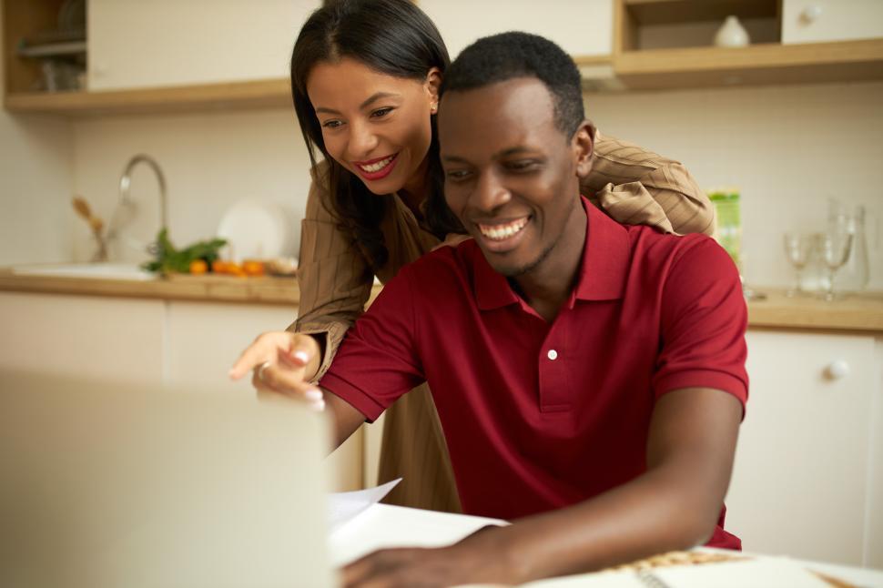 Free Image of Happy ethnicity couple at kitchen browsing online shops on laptop, smiling, dark-skinned elegant charming woman pointing at screen with cheerful face expression, african man typing with joyful look 