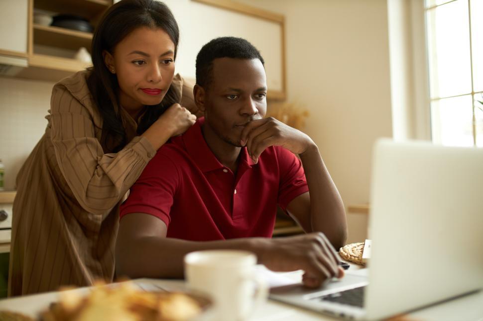 Free Image of Concentrated young adult husband and wife looking at screen of modern laptop, reading article, surfing real estate or traveling websites, woman leaning on man s back, sitting at table in kitchen 