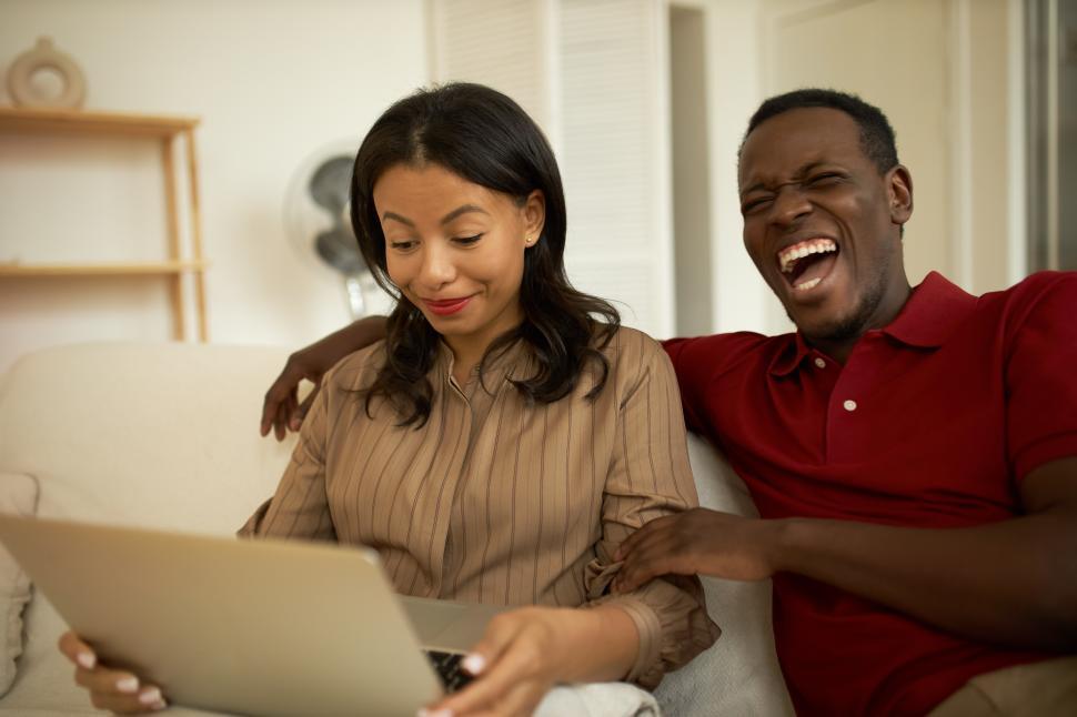 Free Image of Multiracial couple spending leisure time together watching show or funny video on laptop, man in red t-shirt laughing out loud sitting next to his smiling elegant pretty girlfriend on couch 