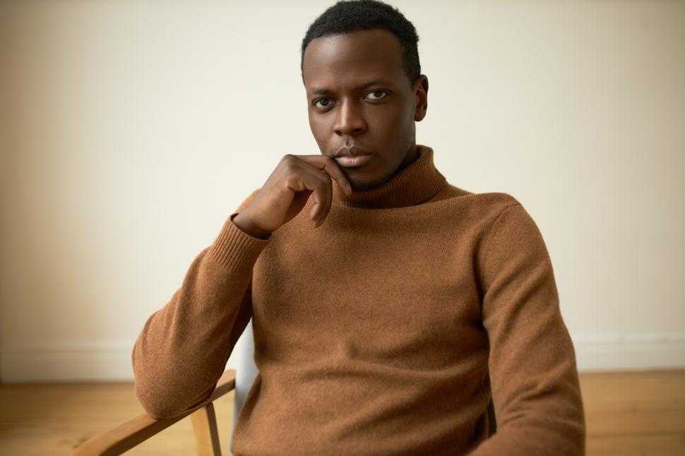 Free Image of Studio portrait of handsome brutal serious and confident African American man in brown polo neck cashmere jumper, looking at camera with thoughtful face expression, resting chin on hand 