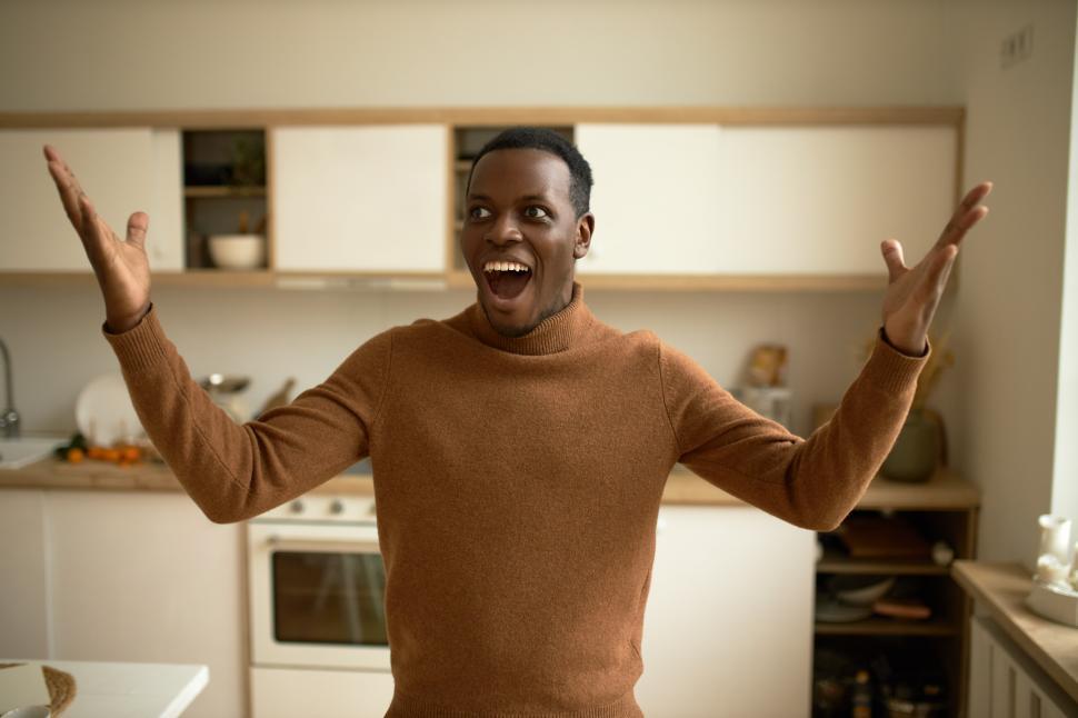 Free Image of Cheerful African American guy in brown polo neck sweater putting hands up, screaming with happiness, making eyes big, standing against kitchen, celebrating moving in his own apartment 