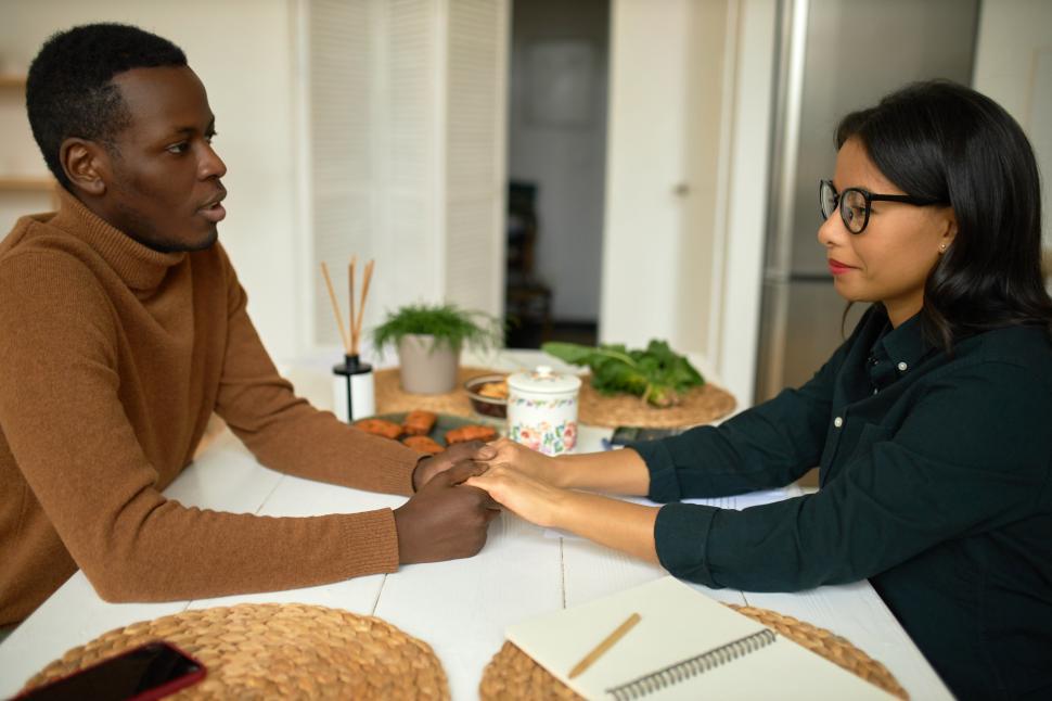 Free Image of Handsome stylish 25-years-old man in turtleneck cashmere jumper sitting next to his dark-skinned pretty young girlfriend in glasses at kitchen table, looking in eyes, proposing her to marry him 