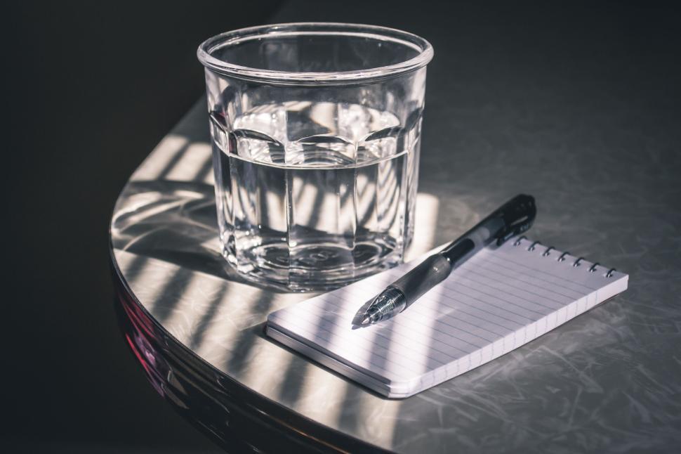 Free Image of Glass of water with pen and notebook on a sunlit table 