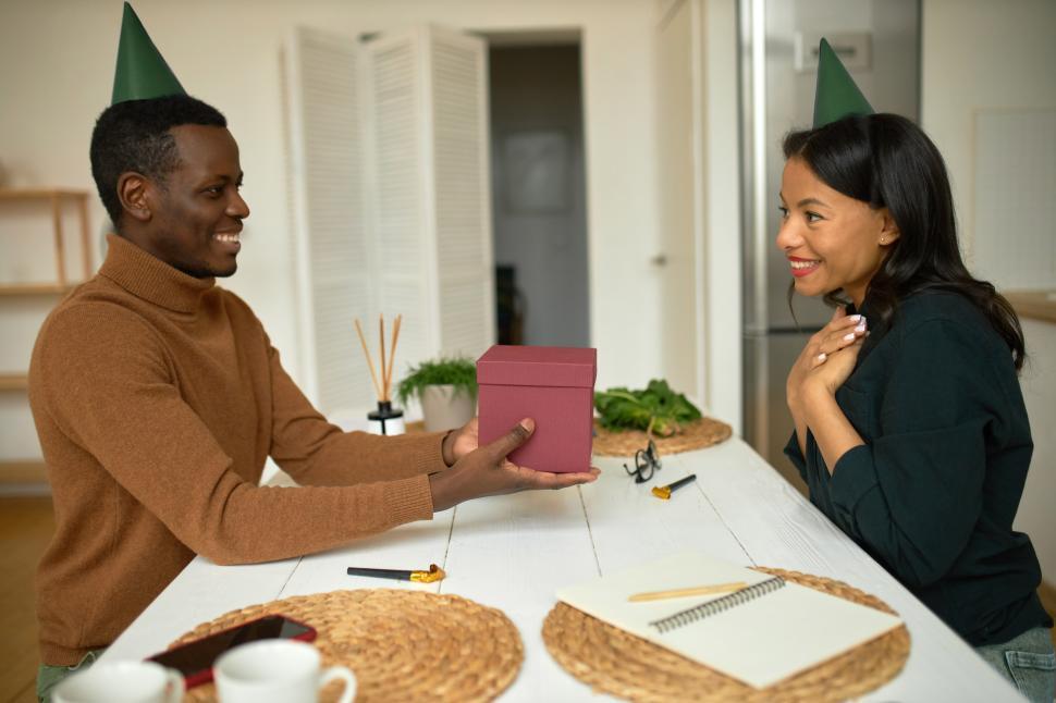 Free Image of Happy birthday. Two lovers sitting at kitchen table in green cones, African man giving present in pink box to his fiance, dark-skinned pretty stylish grateful woman putting hands on chest, smiling 