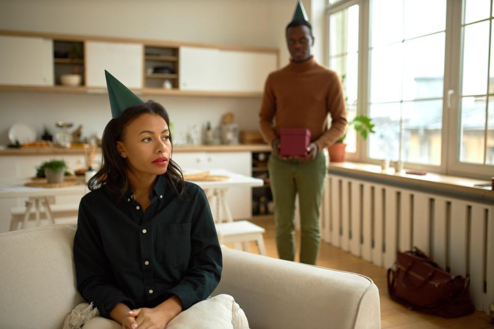 Free Image of Sad birthday party. Upset beautiful woman wearing green shirt and cone on head sitting on sofa in living room, her afro man standing behind with present box trying to make up quarrel 