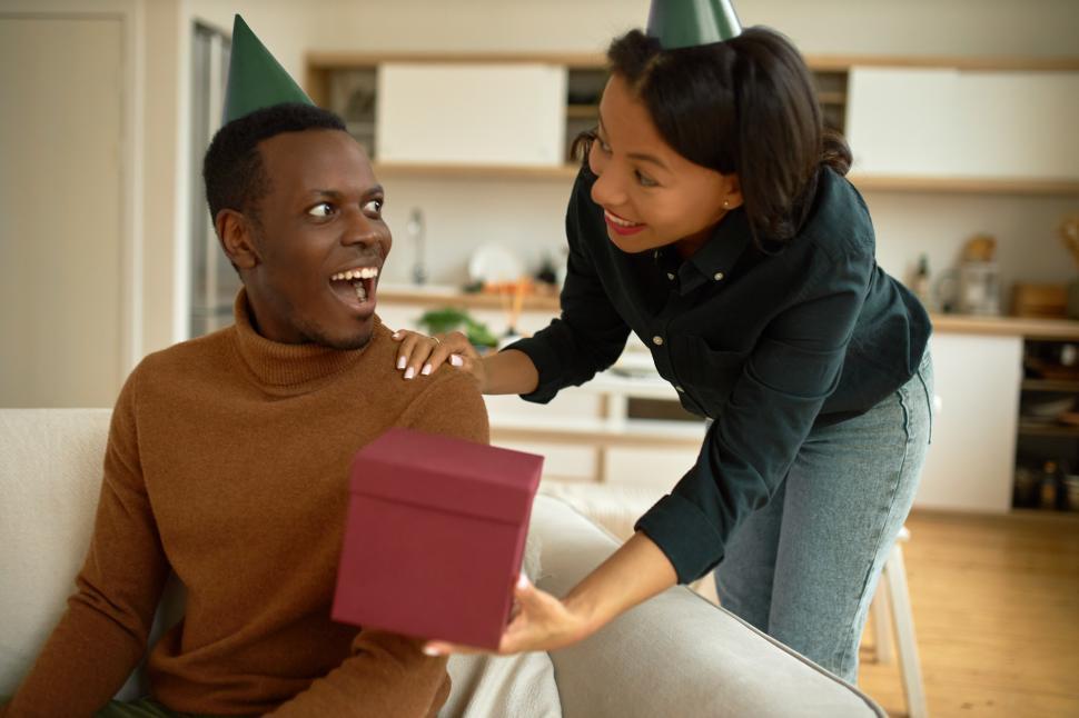 Free Image of Loving and caring charming ethnicity casually dressed stylish woman giving her excited cheerful African boyfriend pink box with gift inside as unexpected surprise, both wearing green birthday cones 