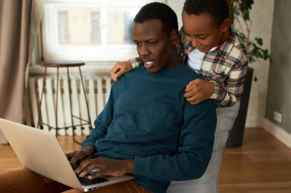 Free Image of Curious African American 8-years-old boy peering over father’s shoulder to see what dad doing on laptop while working online sitting in chair in living-room, tired of waiting for him to play together 