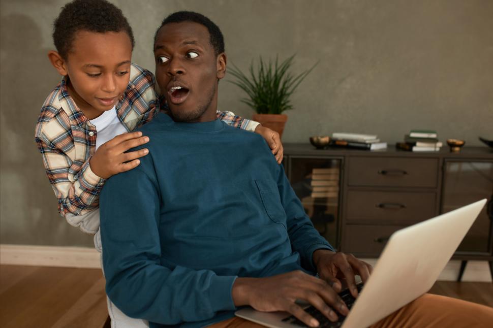 Free Image of Dark-skinned male in blue jumper sitting in chair with shocked face noticing naughty African boy in stylish shirt peering over father s shoulder to see his laptop screen with inappropriate content 