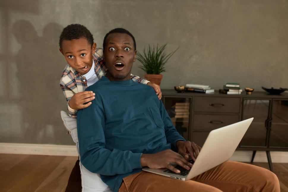 Free Image of Funny 6-years-old boy making oops grimace after seeing forbidden content on fathers laptop screen he watching sitting in living-room after work day, African man looking at camera with shock face 