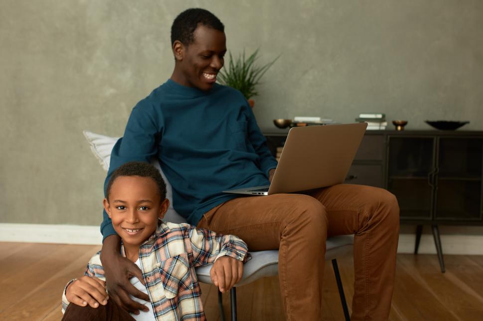 Free Image of Stylish African American dad putting his hand on shoulder of his son playing around on floor while working online, holding laptop on his knees, both feeling joy and happiness. Human relationships 