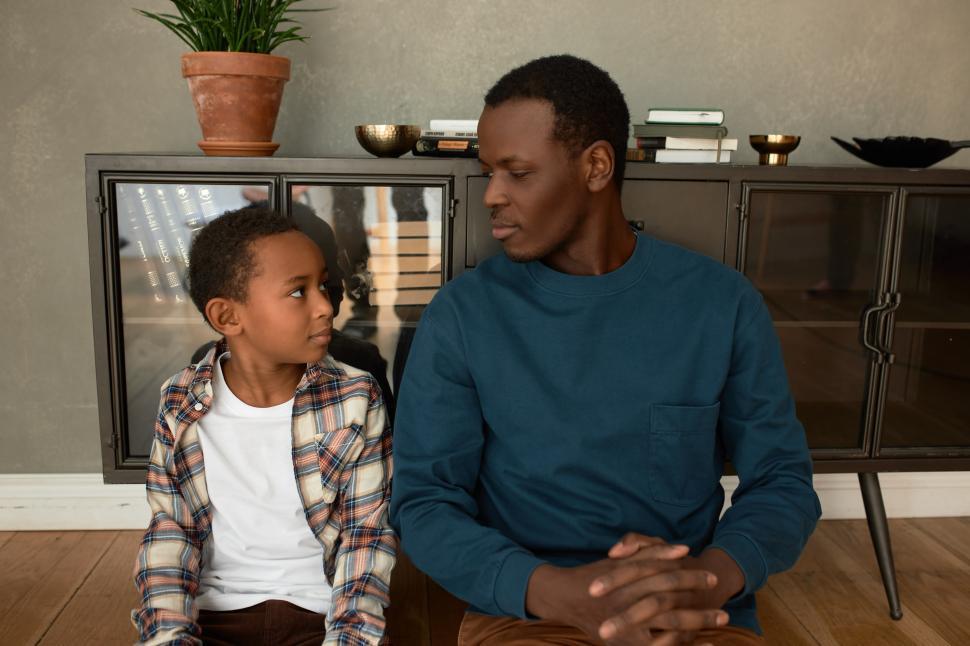 Free Image of Father and son relationships. Male kid and his dad looking at each other with happy calm face after having long serious conversation about child s misbehavior, both leaning on cupboard 