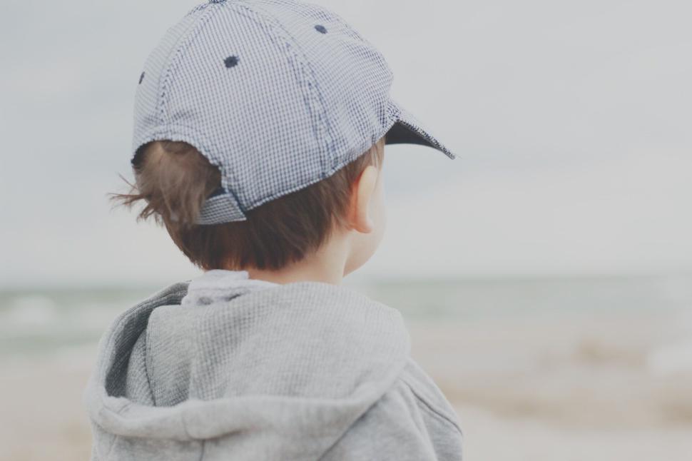 Free Image of Child with cap facing the beach in a grey hoodie 