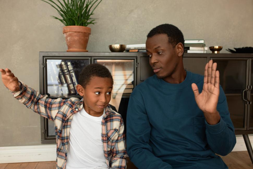 Free Image of Cute little preschool boy in plaid shirt high-fiving to his dad after striking deal, looking positive, having fun sitting on floor against small bookcase. Father and son relationships 