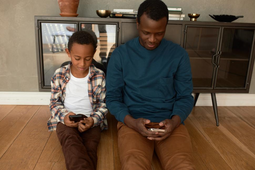 Free Image of Communication gap. Dark-skinned father and his son sitting on floor in living-room and using smart phones, in silence, not noticing each other, having Internet and e-games addictions 