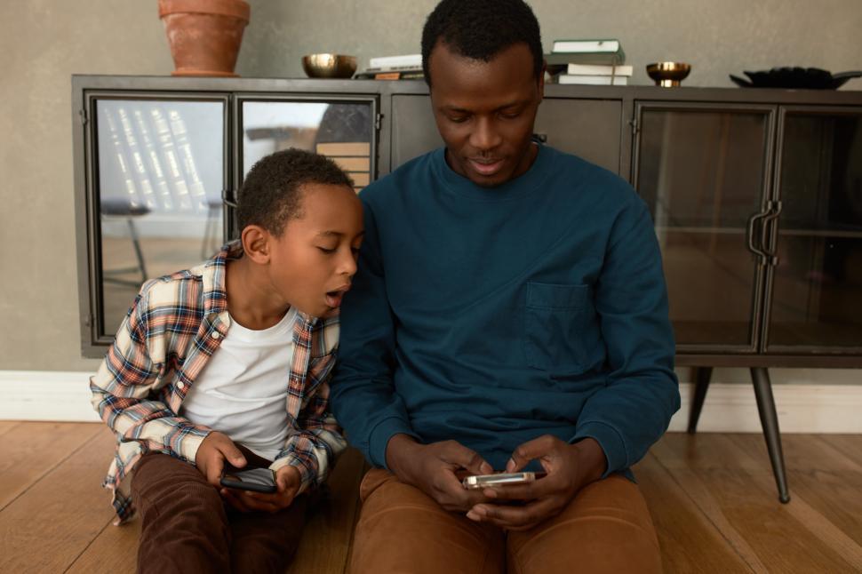 Free Image of Cute black ethnicity male child in stylish casual clothes sitting on floor with mobile phone next to his dad, looking at his gadget to learn rules of game he playing. Father and son relationships 