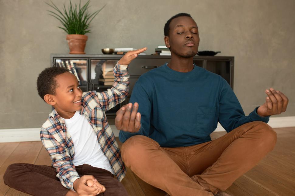 Free Image of Smiling cheerful naughty brown-skinned casually dressed boy of 8-years-old disturbing his dad meditating in lotus posture, opening one eye, to catch little prankster. Human relationships 
