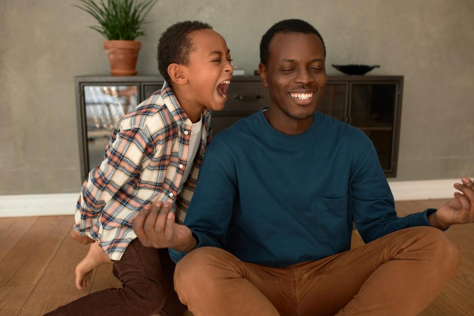 Free Image of African American laughing dad trying to meditate and keep calm while his misbehaving dark-skinned 9-years-old son screaming in his ear with frustration, demanding absolute attention 