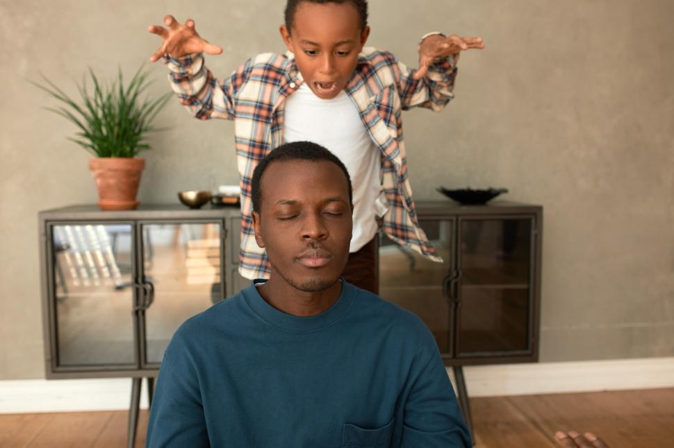 Free Image of Naughty male child pretending tiger standing in attacking posture behind his handsome dark-skinned father sitting on floor with closed eyes after returning from work, having short nap or relaxing 