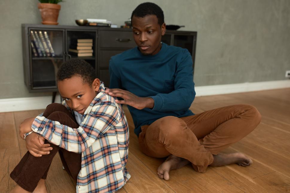 Free Image of Angry little African American boy sitting in closed position with frustration on his face while his dad trying to support him, putting hand on his shoulder, both sitting on floor against living-room 