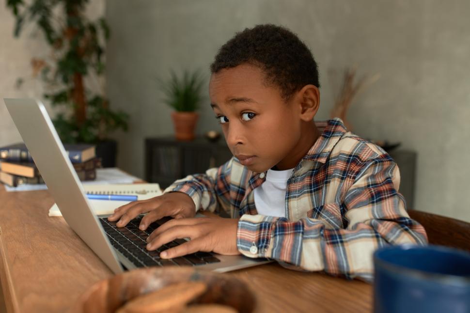 Free Image of Portrait of little African American boy in trendy shirt doing sneaky things, reaching his father s laptop, looking at camera with scared, canny face expression. Children misbehaving concept 