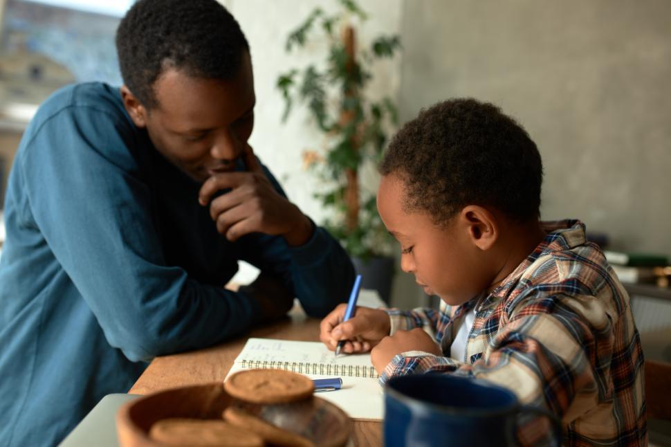 Free Image of Diligent cute little boy with dark skin writing down numbers or letters in his copy book, sitting next to his proud father tutoring him, near cookies and cup of tea. Parenting. School preparation 