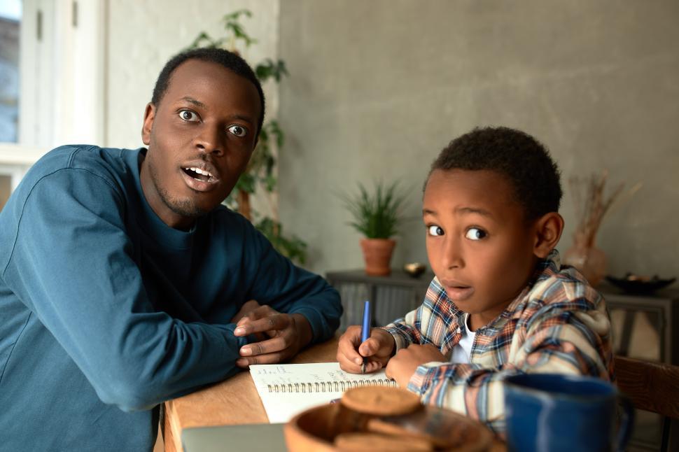 Free Image of Indoor picture of dark-skinned male father sitting with his son at desk in living-room doing homework, having shocked face expressions, scared of exam failure, plate with cookies and mug on table 
