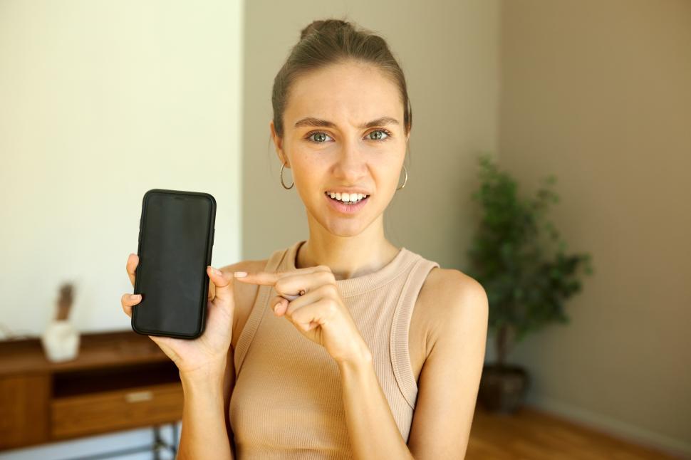 Free Image of Girl with awkward facial expression pointing at black screen of her smartphone, frowning with confusion, having troubles with mobile application or wireless internet connection standing in living room 