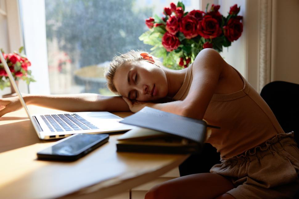 Free Image of Portrait of adorable tired exhausted student girl sleeping on table in front of laptop, smartphone and copy book after studying online, having rest sitting near window with red roses in vases 
