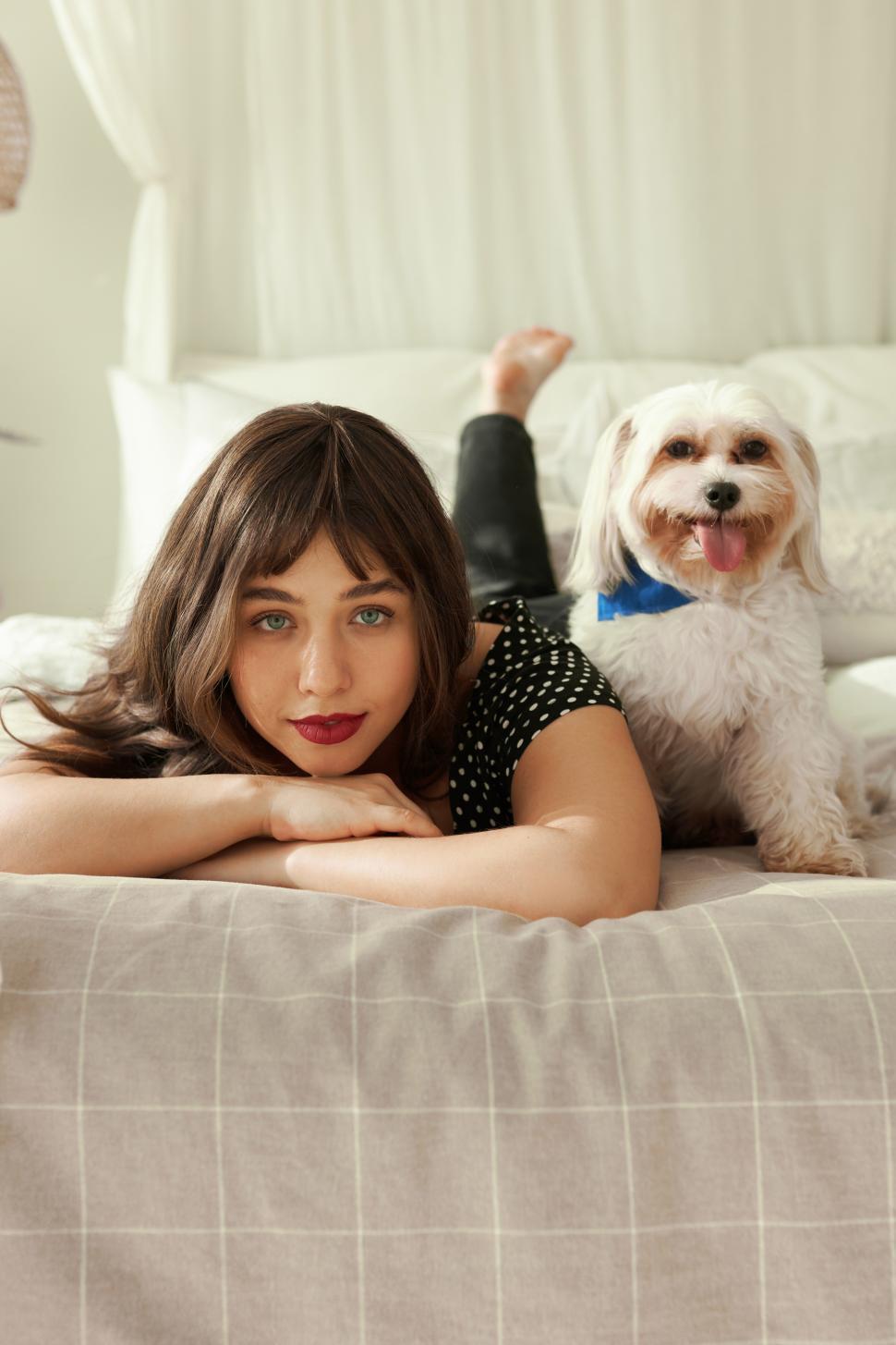 Free Image of Vertical portrait of pretty charming female with red lipstick on lips lying on bed with her white chinese dog pet next to her, looking at camera resting chin on hands, spending weekend at home 