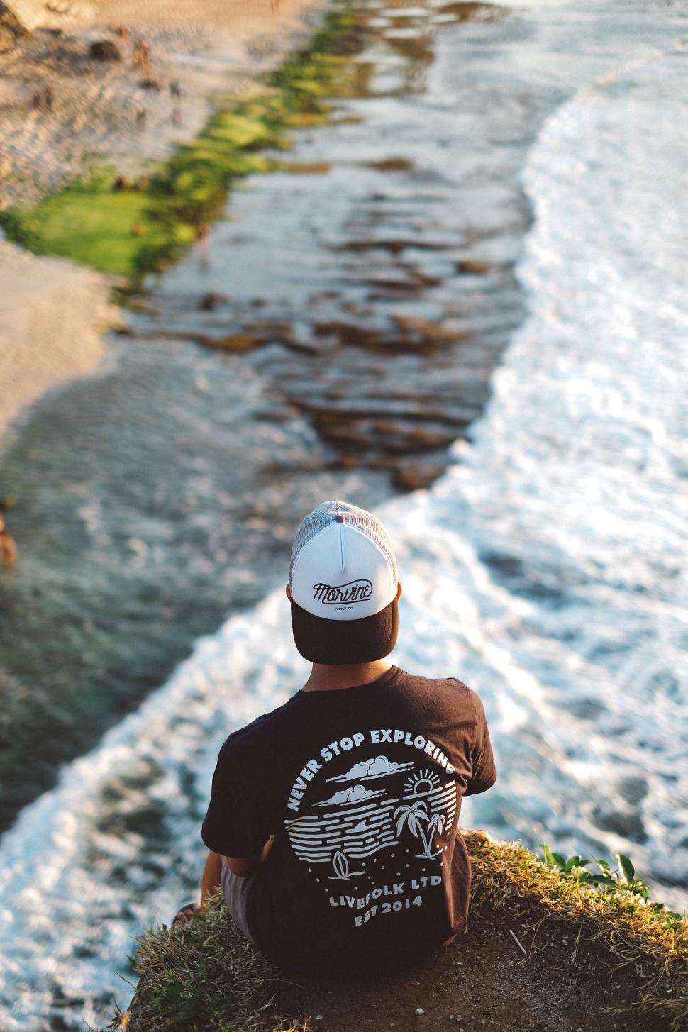 Free Image of Person wearing cap sitting cliffside near water edge 