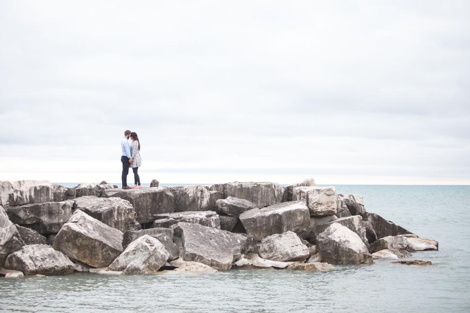 Free Image of Couple standing on large rocks by the shore of ocean 