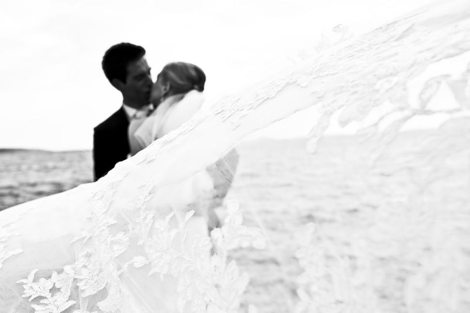 Free Image of A couple embracing under a flowy veil by the water. 