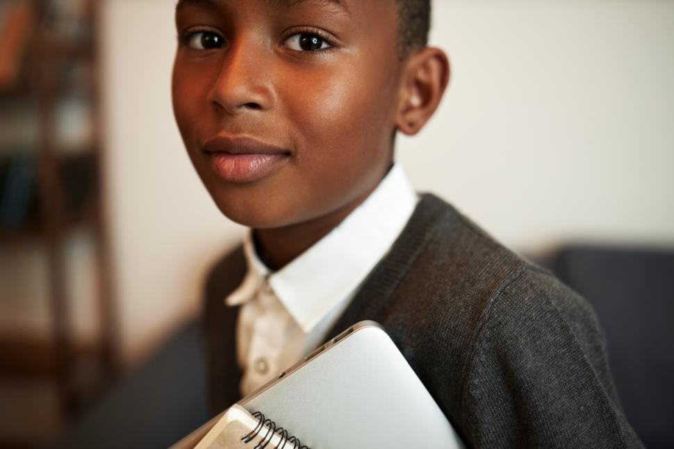 Free Image of Closeup cropped image of black teen schoolboy on his way for class at school, standing on blurred background of classroom interior, holding copybook and laptop for interactive lessons in hands 
