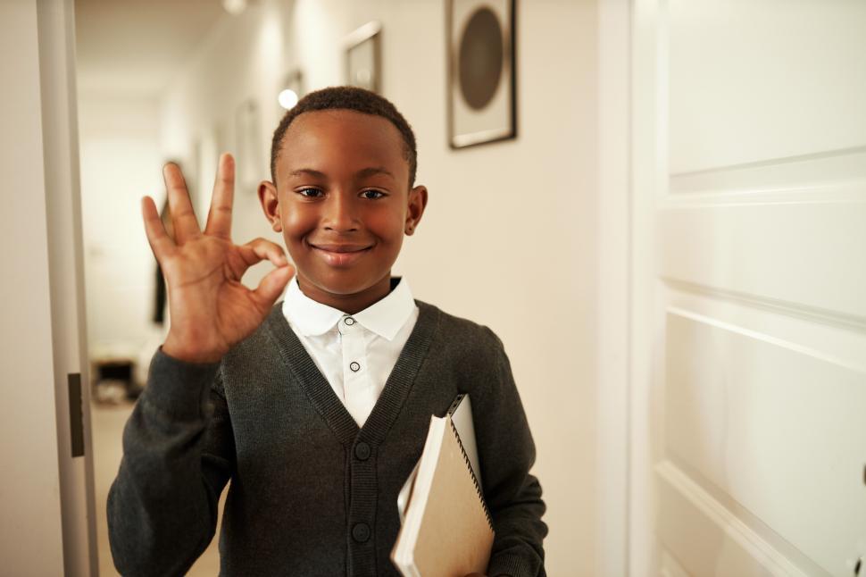 Free Image of Brilliant successful african american school kid with copybook in hands showing ok sign with fingers standing in doorway, sharing good news with parents after passing exam and getting excellent mark 
