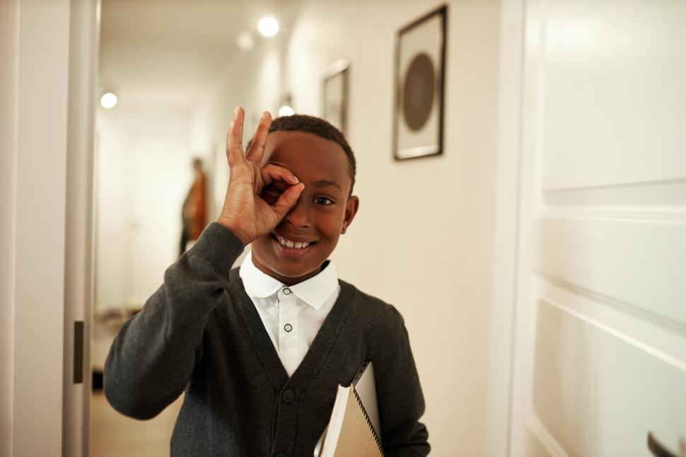 Free Image of Black male schoolkid making faces, putting ok shaped fingers to right eye and looking through at camera with crazy smile, overjoyed with good marks and finishing academic year successfully 