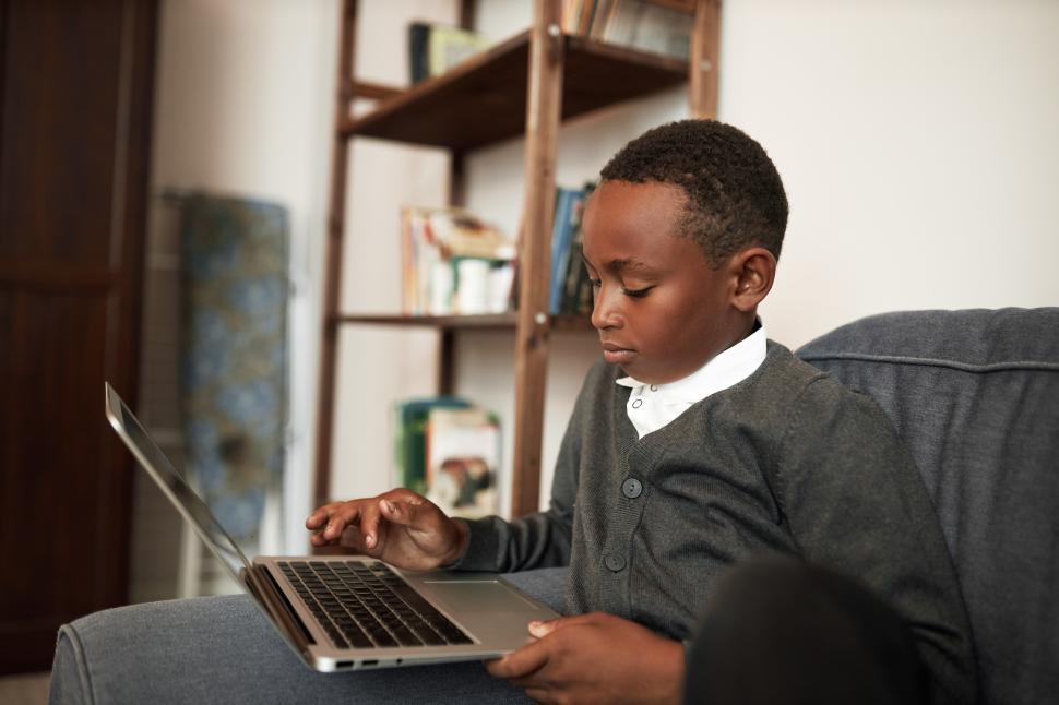 Free Image of Homeschooling concept. Side view of concentrated african american schoolboy in cardigan and white shirt using laptop, entering right variant passing test online sitting on cozy sofa in his room 