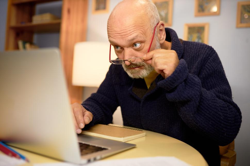Free Image of Aging and eyesight problems. Side view image of senior bald bearded grandfather in elegant stylish blue cardigan using laptop in his cabinet, reading article, adjusting glasses on his nose 