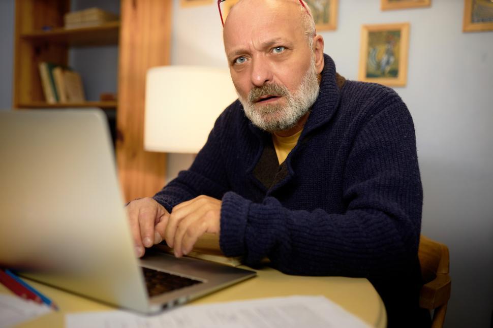 Free Image of Side view of thoughtful senior novelist in elegant cardigan creating new plot of his novel, sitting in his cabinet at table in front of laptop, typing, looking at camera with thoughtful face 
