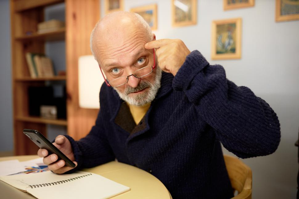 Free Image of Stressed senior financial trader in cardigan and glasses looking at camera putting index finger to temple encourage you to think better before buying or selling crypto currency, holding phone at table 